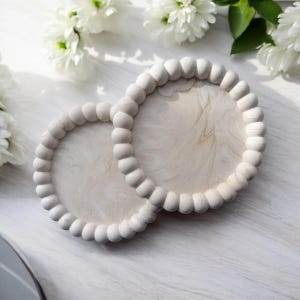 Puede incluir: Dos bandejas redondas de madera blanca con un borde de cuentas. Las bandejas están anidadas y tienen un diseño de mármol.