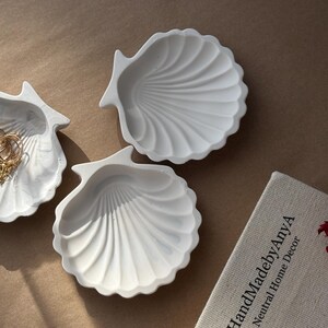 May include: Three white ceramic seashell-shaped dishes. The dishes are arranged on a beige surface. One dish has a gold chain inside.