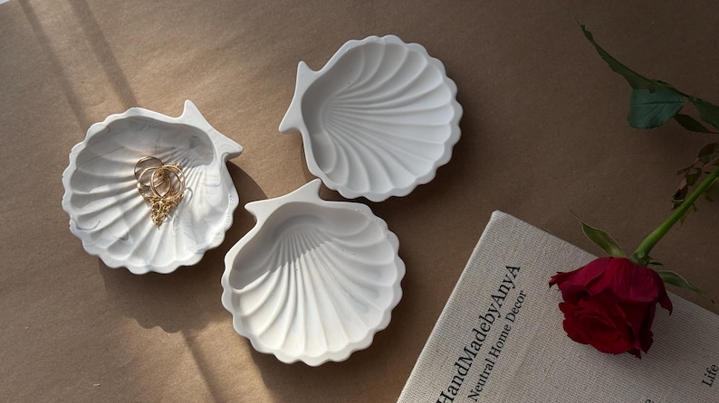 May include: Three white ceramic seashell-shaped dishes. The dishes are arranged on a beige surface. One dish has a gold chain inside.