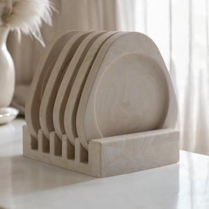 Puede incluir: Un juego de seis posavasos de cerámica de color blanco roto con forma redondeada y ligeramente curvada. Los posavasos están apilados en un soporte del mismo material.