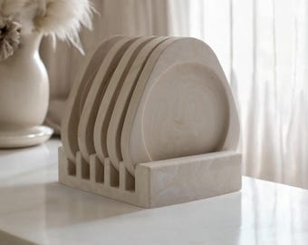 May include: A set of six off-white ceramic coasters with a rounded, slightly curved shape. The coasters are stacked in a holder made of the same material.