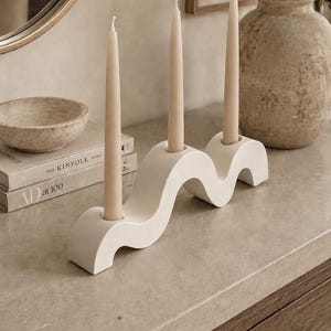 Op de afbeelding: Een witte golvende kandelaar met drie ronde openingen, elk met een lange, slanke, beige dinerkaars. De kandelaar heeft een gladde, matte afwerking en een doorlopende golvende vorm die plat op een oppervlak rust.