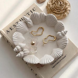 Könnte beinhalten: Ästhetische Präsentation mit einem weißen muschelförmigen Tablett mit goldfarbenen Ohrringen und einem transparenten blütenförmigen Parfümflakon. Das Tablett liegt auf einem Buch mit dem Wort "AESTHETIC". Zusätzliche Muscheln und eine dekorative Blume.