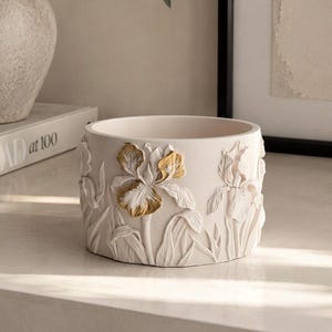 Op de afbeelding: Een cilindervormige, gebroken witte plantenbak met reliëfsculpturen van irissen rondom de buitenkant. Eén opvallende irisbloem is geaccentueerd met metallic goudkleurige verf op de bloemblaadjes, terwijl de overige bloemmotieven mat wit zijn.