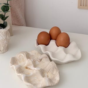 May include: Two ceramic egg holders and a decorative dish. One egg holder is white and holds three brown eggs. The other dish has a marbled beige and white pattern. The items are on a white surface.