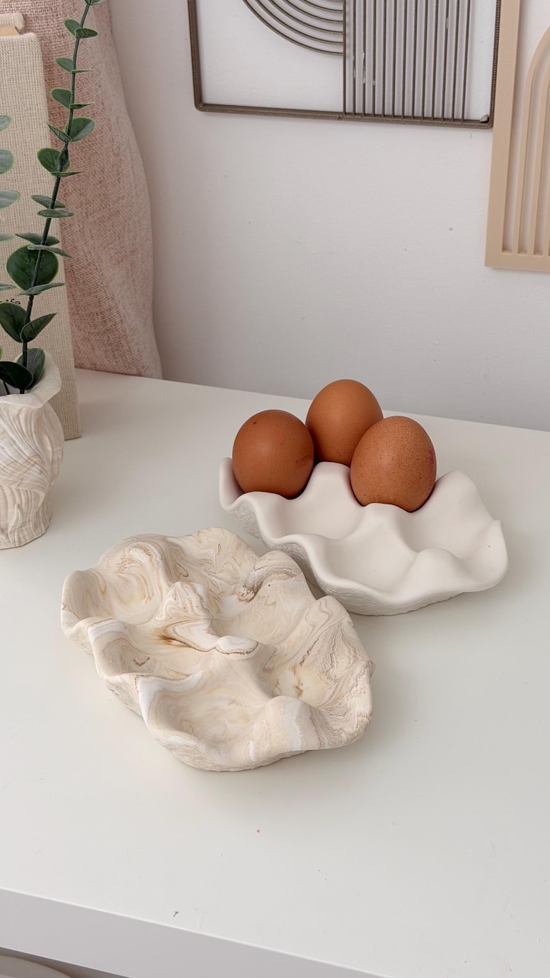 May include: Two ceramic egg holders and a decorative dish. One egg holder is white and holds three brown eggs. The other dish has a marbled beige and white pattern. The items are on a white surface.