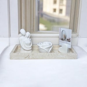 Op de afbeelding: Een witte keramische dienblad met een marmerpatroon, met een witte keramische sculptuur van een koppel dat elkaar omarmt, een witte keramische roos en een wit hartvormig fotolijstje met een foto van een kamer met een spiegel en een plant.