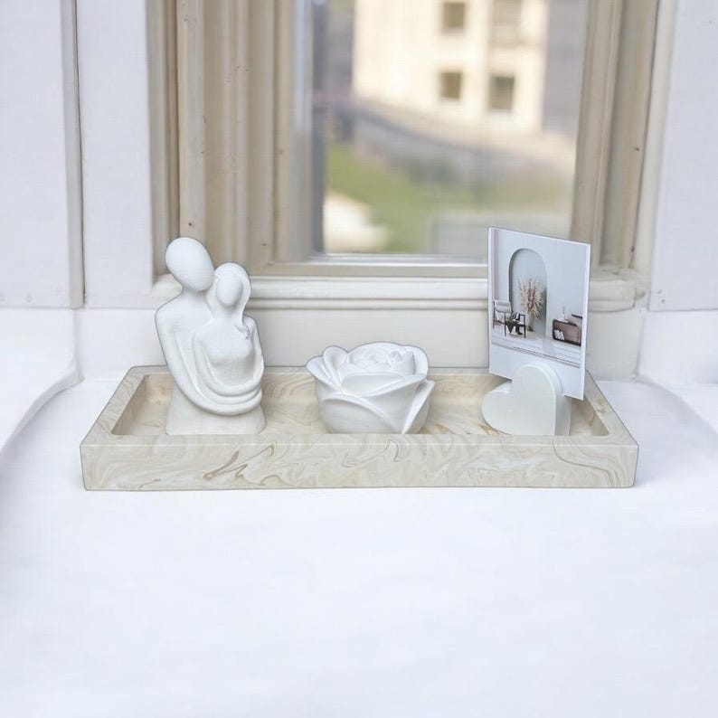 Op de afbeelding: Een witte keramische dienblad met een marmerpatroon, met een witte keramische sculptuur van een koppel dat elkaar omarmt, een witte keramische roos en een wit hartvormig fotolijstje met een foto van een kamer met een spiegel en een plant.