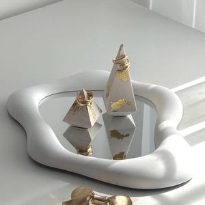 May include: A white, organically shaped tray with a mirrored surface features two pyramid ring holders. The light grey ring holders have gold leaf details and gold rings. A gold bow-shaped jewellery piece sits in front of the tray.