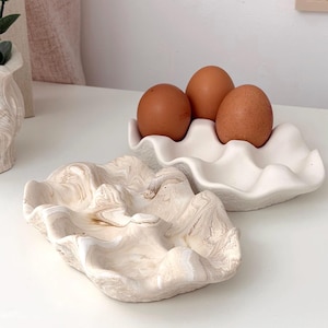 May include: Two ceramic egg holders. One is white and holds three brown eggs. The other is a marbled beige and white clam shell design. Both are on a white surface.