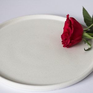 May include: A white ceramic plate with a single red rose on top. The rose is partially off the plate and has green leaves.