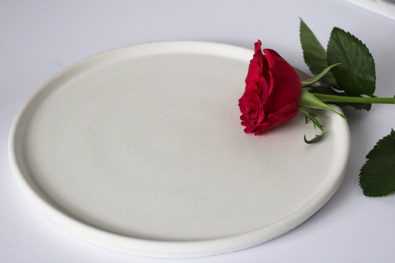 May include: A white ceramic plate with a single red rose on top. The rose is partially off the plate and has green leaves.