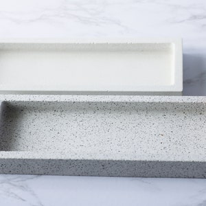 Op de afbeelding: Twee rechthoekige dienbladen, een wit en een met een gespikkelde grijze afwerking. De dienbladen zijn gemaakt van een glad, getextureerd materiaal en zijn perfect om kleine voorwerpen te presenteren of snacks te serveren.