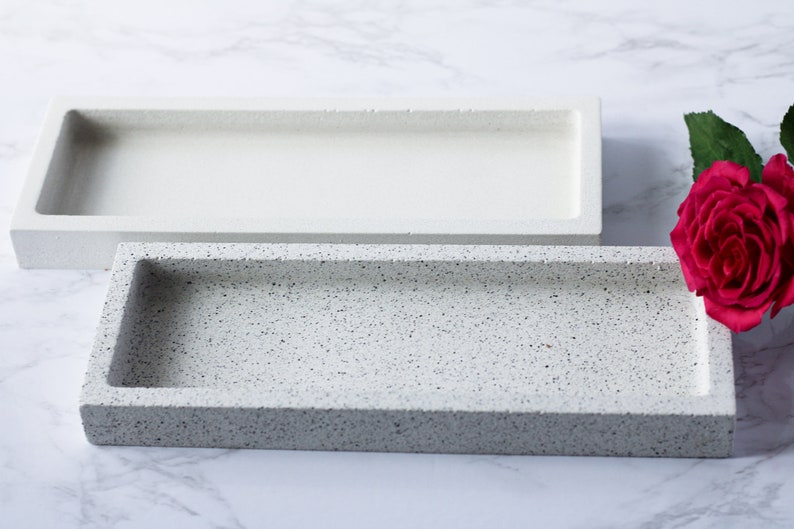 Op de afbeelding: Twee rechthoekige dienbladen, een wit en een met een gespikkelde grijze afwerking. De dienbladen zijn gemaakt van een glad, getextureerd materiaal en zijn perfect om kleine voorwerpen te presenteren of snacks te serveren.