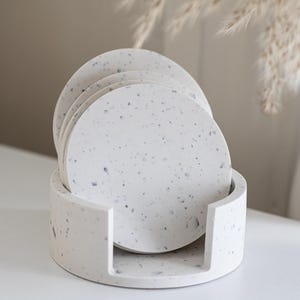 Puede incluir: Un juego de seis posavasos redondos de material blanco moteado, colocados en un soporte a juego. Los posavasos y el soporte tienen un aspecto similar al terrazo con peque&ntilde;as motas oscuras. El dise&ntilde;o es minimalista y moderno, adecuado para la decoraci&oacute;n del hogar.