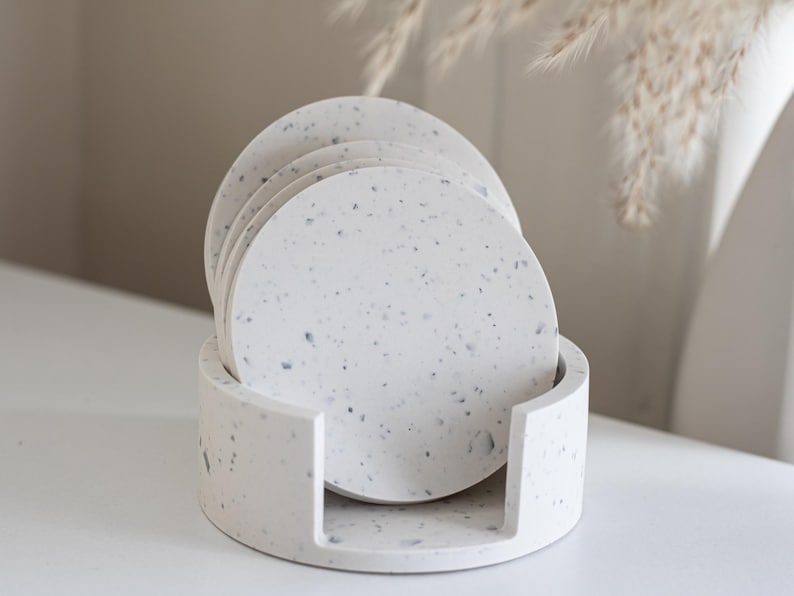 Puede incluir: Un juego de seis posavasos redondos de material blanco moteado, colocados en un soporte a juego. Los posavasos y el soporte tienen un aspecto similar al terrazo con peque&ntilde;as motas oscuras. El dise&ntilde;o es minimalista y moderno, adecuado para la decoraci&oacute;n del hogar.