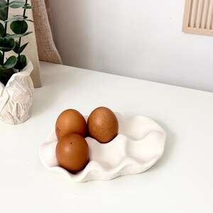 May include: A white ceramic egg holder with a wavy design, holding three brown eggs. The eggs are evenly spaced in the holder. The scene is set on a white surface, with a plant and decorative wall art in the background.