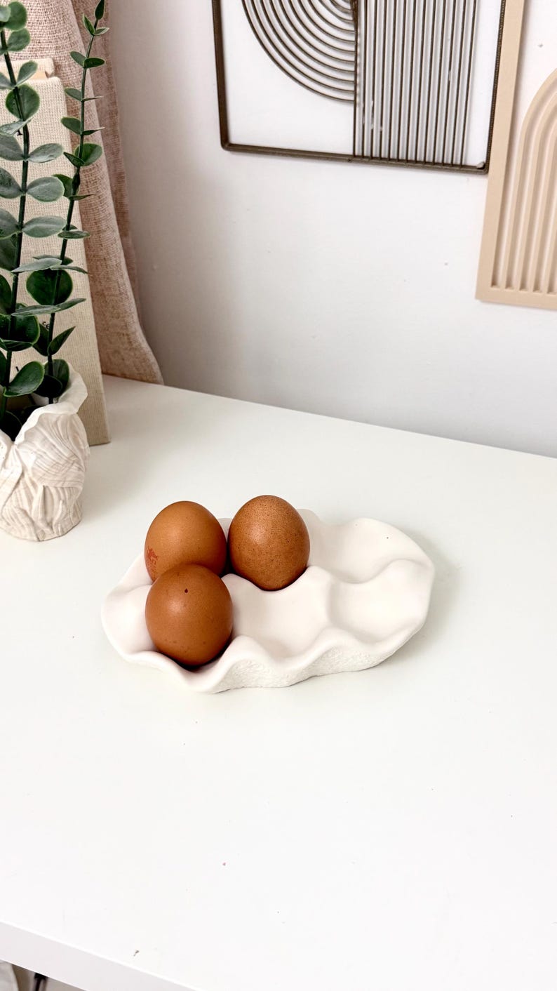 May include: A white ceramic egg holder with a wavy design, holding three brown eggs. The eggs are evenly spaced in the holder. The scene is set on a white surface, with a plant and decorative wall art in the background.