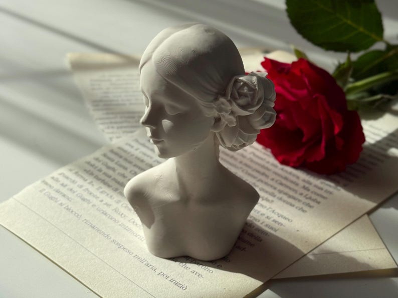 Puede incluir: Busto de mujer de yeso blanco con una flor en el pelo. El busto est&aacute; sentado en un libro con una rosa roja en el fondo.