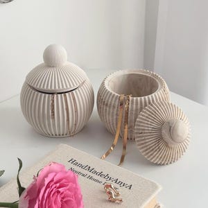 Könnte beinhalten: Zwei cremefarbene, gerippte Keramikgläser mit Deckeln, eines offen, mit Goldschmuck. Ein Buch mit dem Text "HandMadebyAnyA" und eine rosa Rose befinden sich im Vordergrund. Die Gläser stehen auf einer weißen Oberfläche.