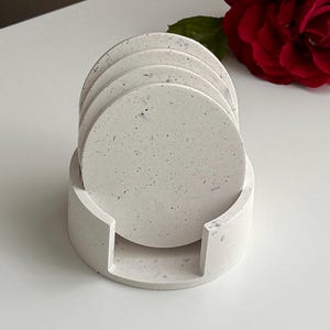 Könnte beinhalten: Ein Set aus vier runden, gesprenkelten, hellgrauen Untersetzern in einem passenden Halter. Die Untersetzer und der Halter bestehen aus einem betonartigen Material. Der Halter hat ein gebogenes Design mit einem Schlitz für einfachen Zugriff. Eine rote Rose ist im Hintergrund.