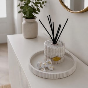 May include: A white marble reed diffuser with black reeds sits on a matching tray, alongside a decorative bow with gold accents. A small vase with greenery is in the background, suggesting home decor.
