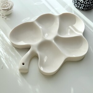 May include: A white ceramic artist's palette with a clover-like shape and multiple wells for mixing paints. The palette has a handle with a small hole. The background includes a white surface and decorative elements.