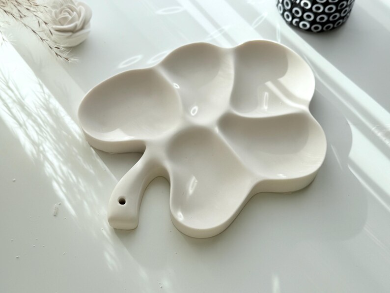 May include: A white ceramic artist's palette with a clover-like shape and multiple wells for mixing paints. The palette has a handle with a small hole. The background includes a white surface and decorative elements.
