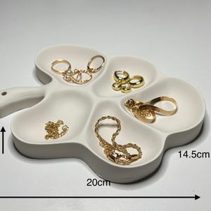 May include: A white, clover-shaped jewelry tray with six compartments. The tray is 20 cm long, 14.5 cm wide, and 2 cm deep. Gold jewelry, including rings, earrings, and necklaces, is displayed in the tray.