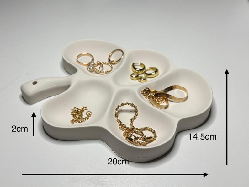 May include: A white, clover-shaped jewelry tray with six compartments. The tray is 20 cm long, 14.5 cm wide, and 2 cm deep. Gold jewelry, including rings, earrings, and necklaces, is displayed in the tray.