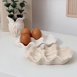 May include: A decorative white ceramic egg holder with three brown eggs. A second, larger, decorative clam shell-shaped dish is in the foreground. A small white vase with greenery and a book with the text "HandMadebyAnyA Home Decor" are in the background.