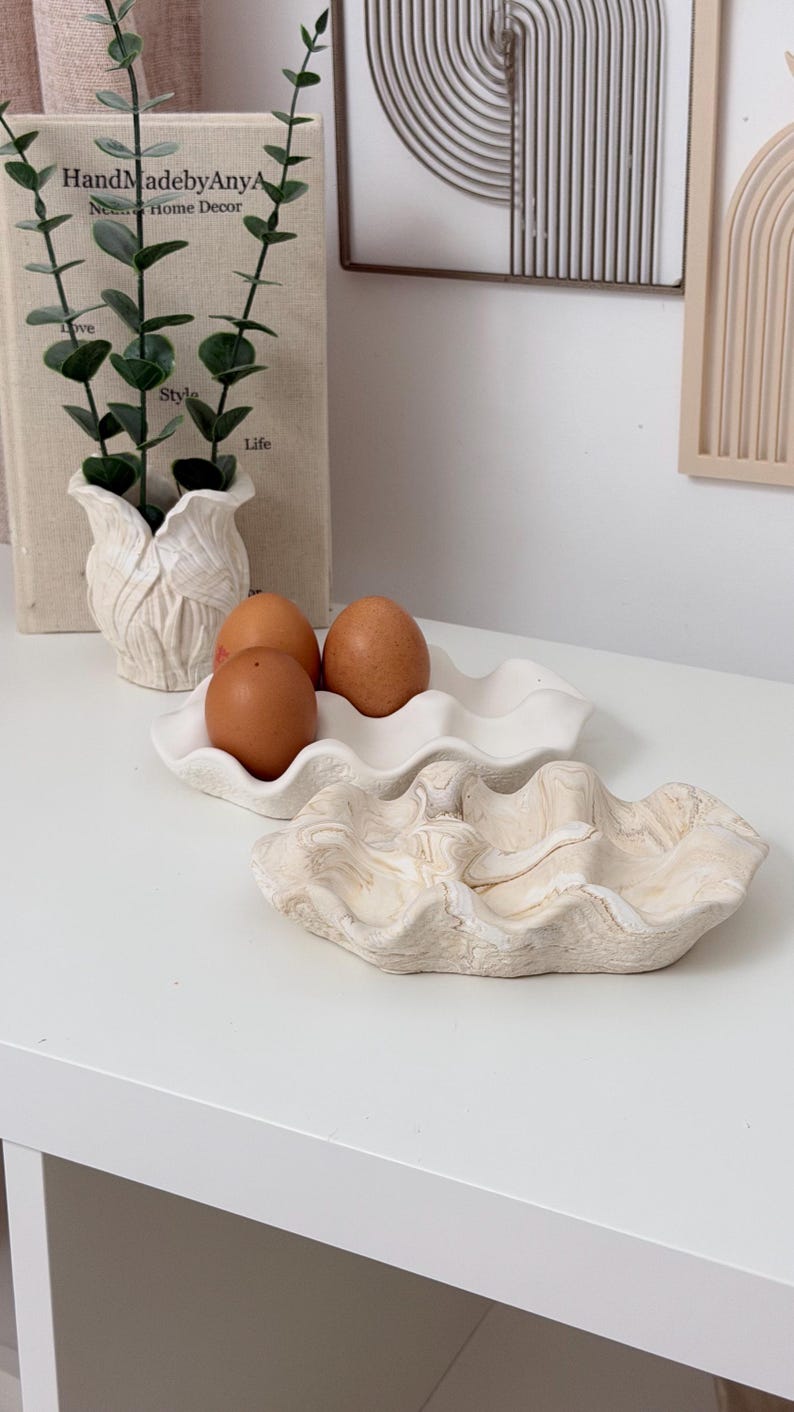 May include: A decorative white ceramic egg holder with three brown eggs. A second, larger, decorative clam shell-shaped dish is in the foreground. A small white vase with greenery and a book with the text "HandMadebyAnyA Home Decor" are in the background.