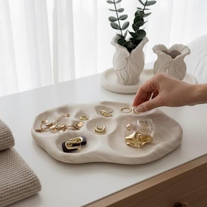 Op de afbeelding: Een crèmekleurige, organisch gevormde sieradenbak met meerdere compartimenten, met gouden ringen, kettingen en een kleine parfumfles. Een hand reikt naar een ring. Twee kleine witte vazen met groen op de achtergrond.