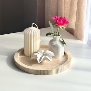 May include: A white wooden tray with a fluted edge, featuring a white candle, a white ceramic vase with a pink rose, and a white ceramic flower.