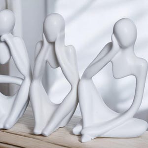 May include: Three white ceramic sculptures of abstract figures in a seated pose. The figures are stylized and have no facial features. They are arranged in a row on a wooden surface.