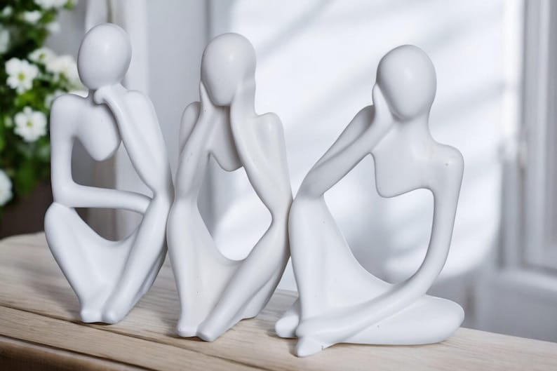 May include: Three white ceramic sculptures of abstract figures in a seated pose. The figures are stylized and have no facial features. They are arranged in a row on a wooden surface.