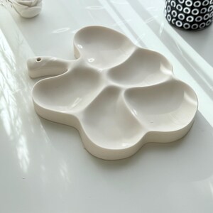 May include: A white ceramic palette with a unique cloud-like shape. The palette features five separate wells for mixing paints and a handle with a small hole. The design is modern and minimalist, perfect for artists.