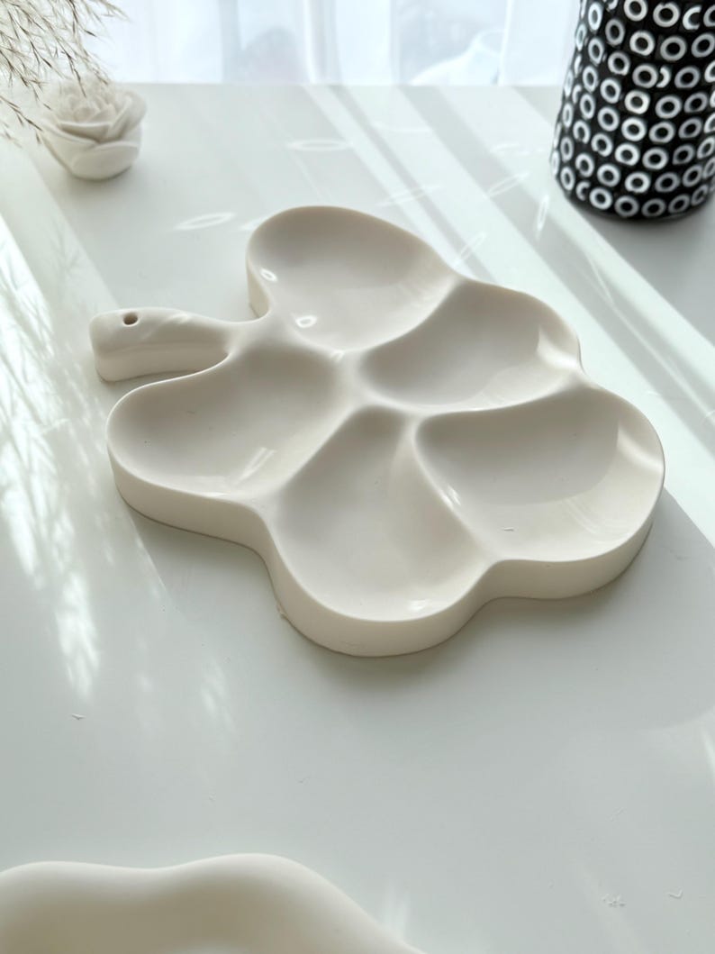 May include: A white ceramic palette with a unique cloud-like shape. The palette features five separate wells for mixing paints and a handle with a small hole. The design is modern and minimalist, perfect for artists.