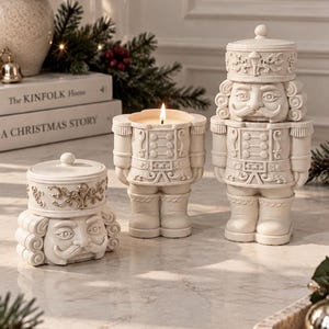 May include: Three cream-coloured nutcracker-shaped containers made of ceramic. One is a lidded jar, one is a candle with a lit wick, and one is a taller lidded jar. Each features detailed moulded facial features, uniforms, and ornate hat designs.
