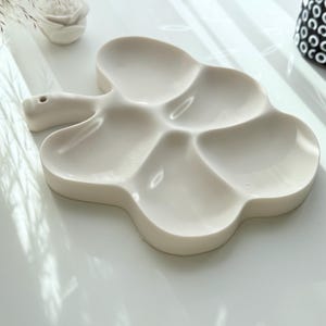 May include: Off-white ceramic paint palette in a clover shape with six sections. The palette has a handle with a small hole. A white ceramic rose and a black and white patterned vase are in the background.