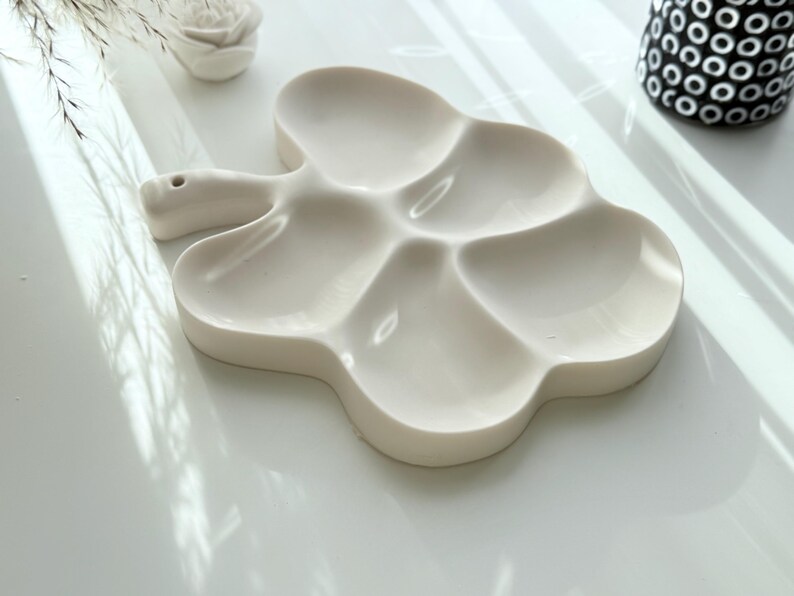 May include: Off-white ceramic paint palette in a clover shape with six sections. The palette has a handle with a small hole. A white ceramic rose and a black and white patterned vase are in the background.