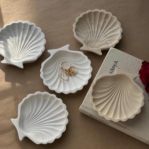 May include: Four white and beige ceramic seashell-shaped trinket dishes. The dishes are arranged on a brown surface with a book titled "Home Decor" visible in the background.