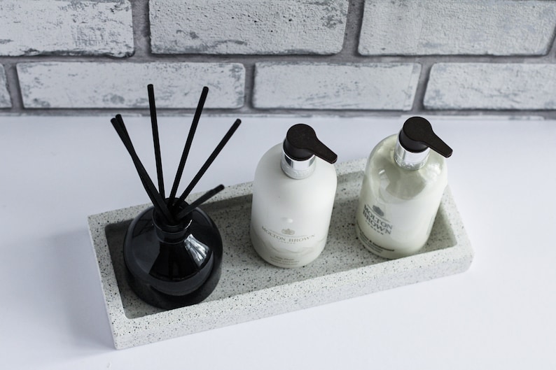 Op de afbeelding: Een wit rechthoekig dienblad met zwarte gespikkelde details bevat twee witte flessen handzeep en een zwarte diffuser met zwarte stokjes. De flessen hebben zwarte pompjes en de tekst "MOLTON BROWN" staat op de voorkant van elke fles.