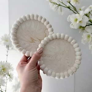May include: Two round, off-white coasters with a beaded edge and a marbled design. The coasters are held in a hand, with white flowers in the background. The coasters are approximately 4 inches in diameter.