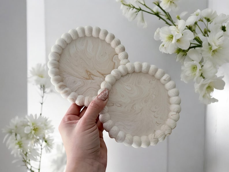 May include: Two round, off-white coasters with a beaded edge and a marbled design. The coasters are held in a hand, with white flowers in the background. The coasters are approximately 4 inches in diameter.
