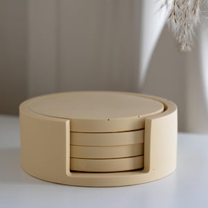 Puede incluir: Un juego de cuatro posavasos redondos beige con un soporte a juego. Los posavasos est&aacute;n apilados dentro del soporte, que tiene un dise&ntilde;o curvo. El juego est&aacute; hecho de un material s&oacute;lido y es adecuado para la decoraci&oacute;n del hogar.