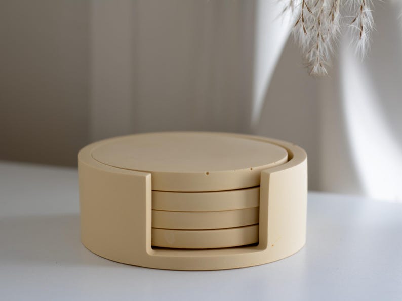 Puede incluir: Un juego de cuatro posavasos redondos beige con un soporte a juego. Los posavasos est&aacute;n apilados dentro del soporte, que tiene un dise&ntilde;o curvo. El juego est&aacute; hecho de un material s&oacute;lido y es adecuado para la decoraci&oacute;n del hogar.