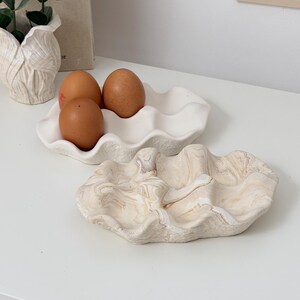 May include: Two decorative ceramic clam shell trays, one white and one with a beige marble design. The white tray holds three brown eggs. The trays are on a white surface, with a neutral home decor theme.