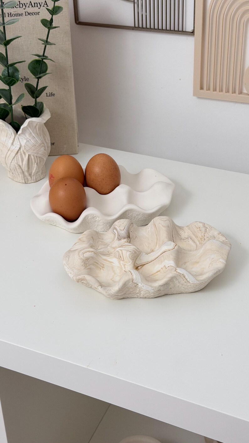 May include: Two decorative ceramic clam shell trays, one white and one with a beige marble design. The white tray holds three brown eggs. The trays are on a white surface, with a neutral home decor theme.