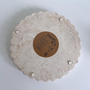 May include: A round, beige Jesmonite tray with a scalloped edge and a marbled pattern. The tray has a brown circular sticker with instructions and care information. Clear, round bumpers are attached to the bottom.